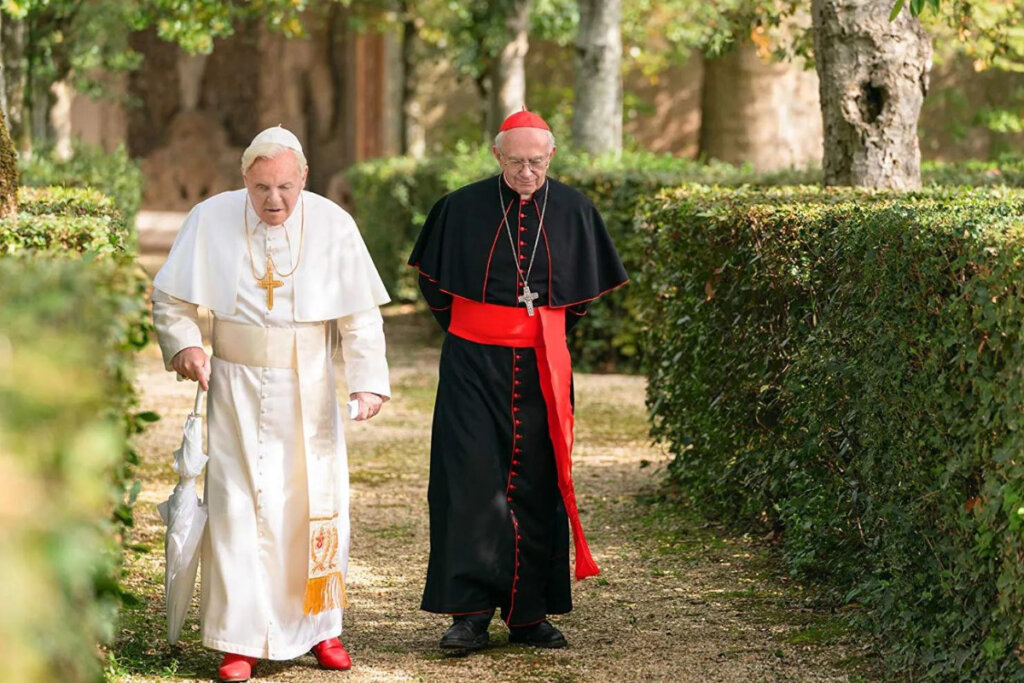 Papa Benedito e Papa Francisco caminhando ao ar livre em cena do filme "Dois Papas"