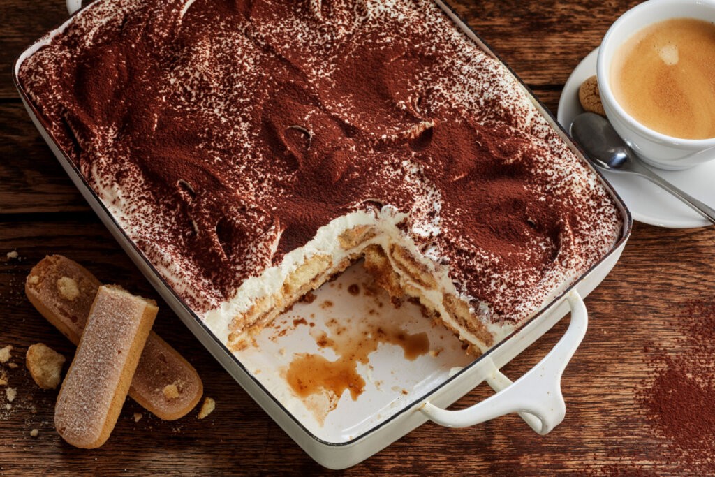 Travessa com tiramisù faltando um pedaço e um pires com xícara de café, colhé e biscoito ao lado